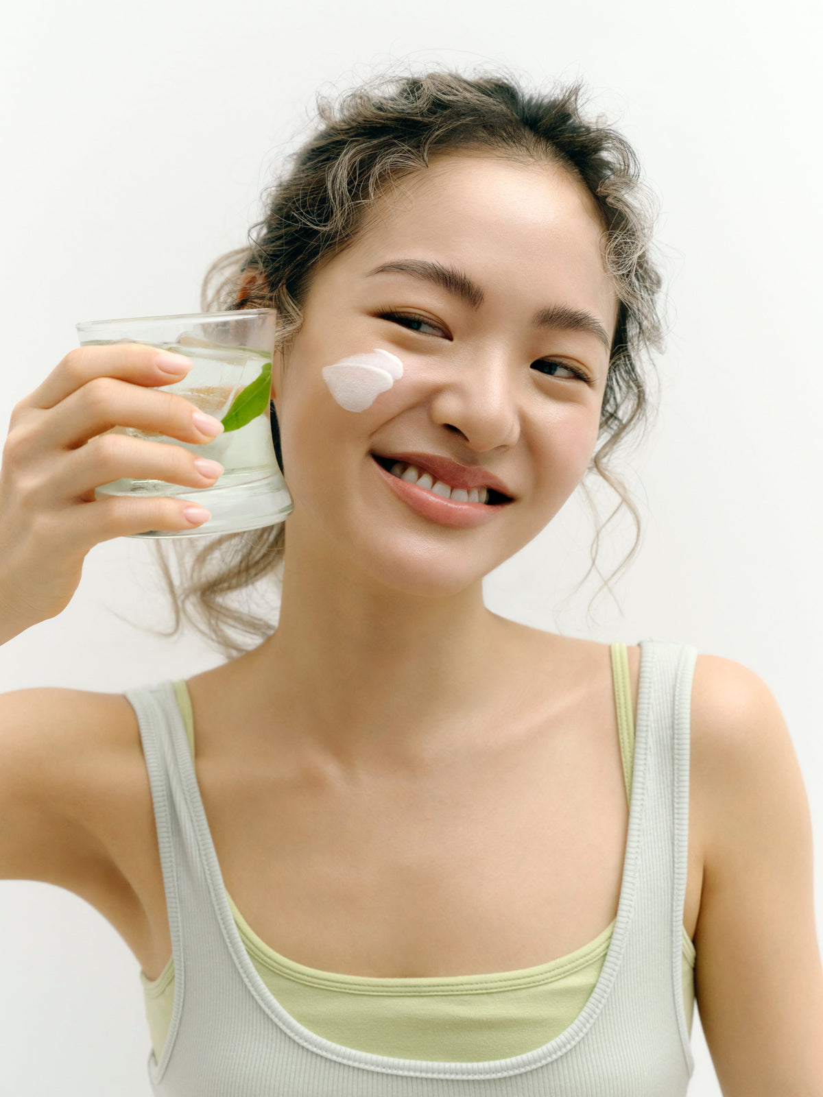 Woman holding a glass of water with a lime, applying cream to her face on a white background