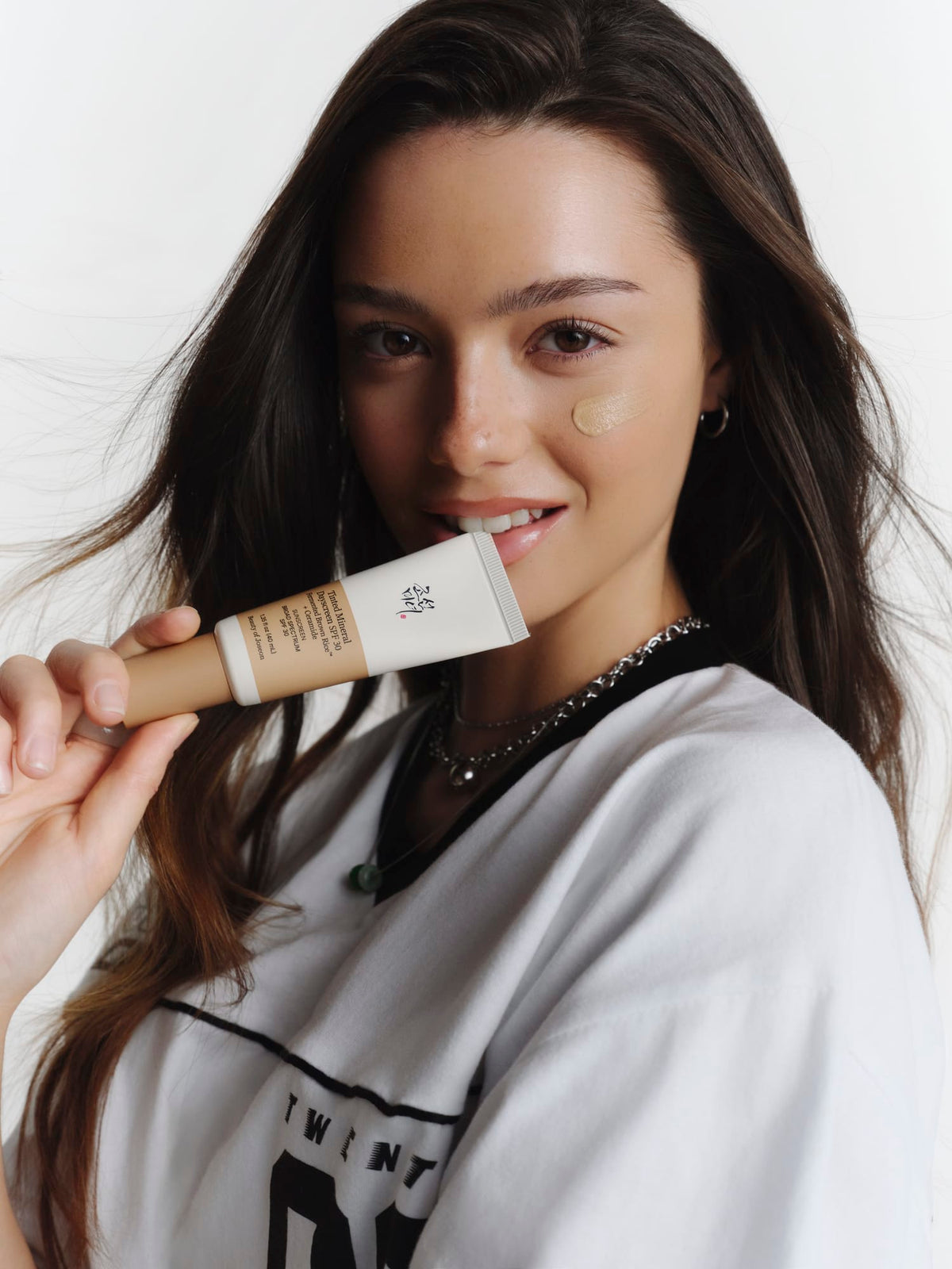 A woman with long dark brown hair holds a Tinted Mineral Dayscreen tube near her mouth, with a dollop of cream foundation on her cheek. She is wearing a white graphic sweatshirt against a clean white background.