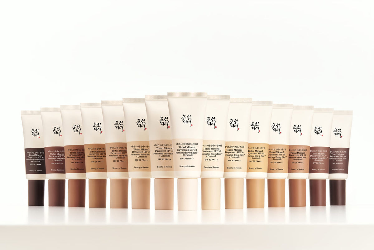 A row of Tinted Mineral Dayscreen tubes in a gradient of skin tones from deep brown to light beige, arranged in a semi-circle on a white reflective surface against a white background.