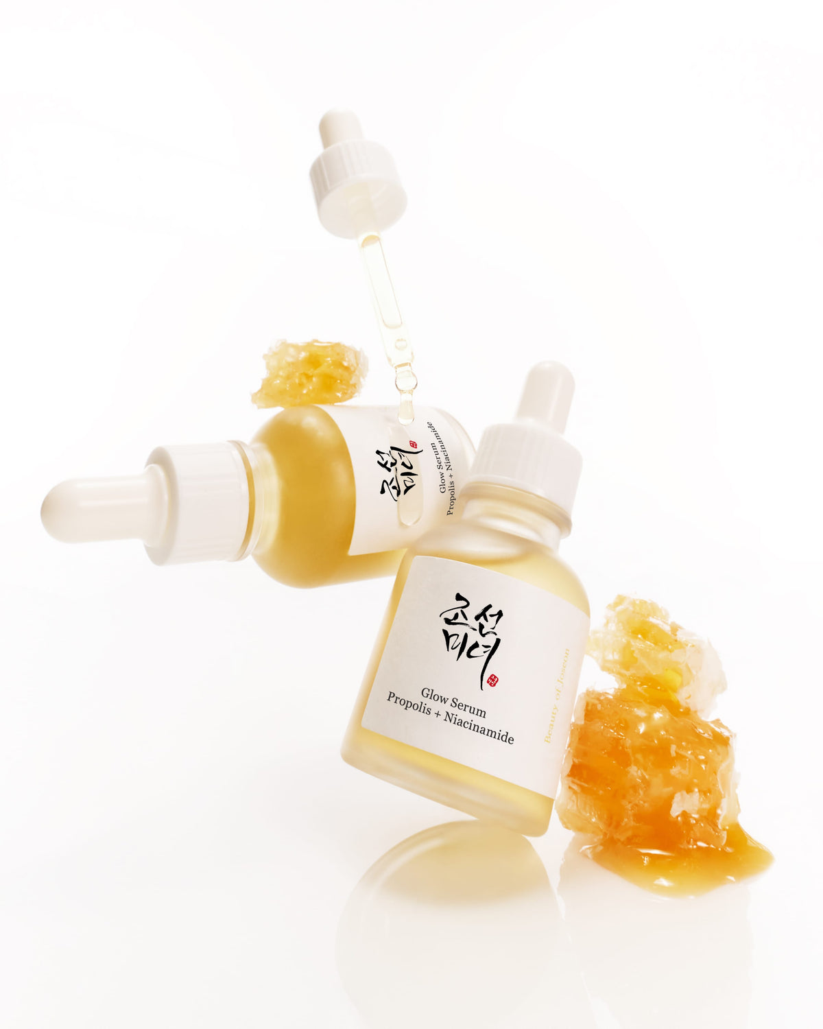 Two small glass dropper bottles of Glow serum arranged on a white reflective surface, with honeycomb pieces and honey dripping into one bottle. The front bottle label reads 'Gluco Serum' and includes Korean script.